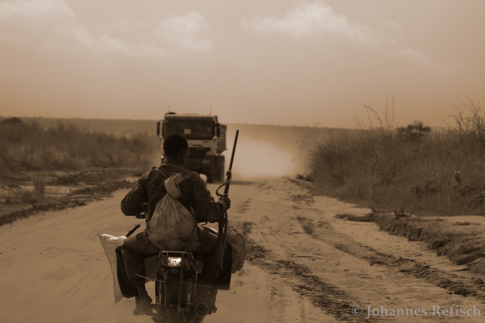 The image shows a bushmeat hunter riding a motorcycle on a dusty road, with a large truck coming towards him. The hunter has a gun in his right hand and is steering with his left. A sack is attached to his back, and others fixed to the side of the motorcycle.